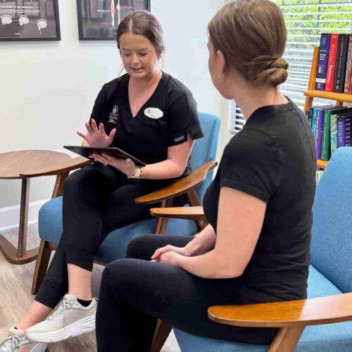 Brittany and a patient sit in armchairs in the waiting area, with Brittany holding a clipboard or tablet as they discuss health history and intake questions.
