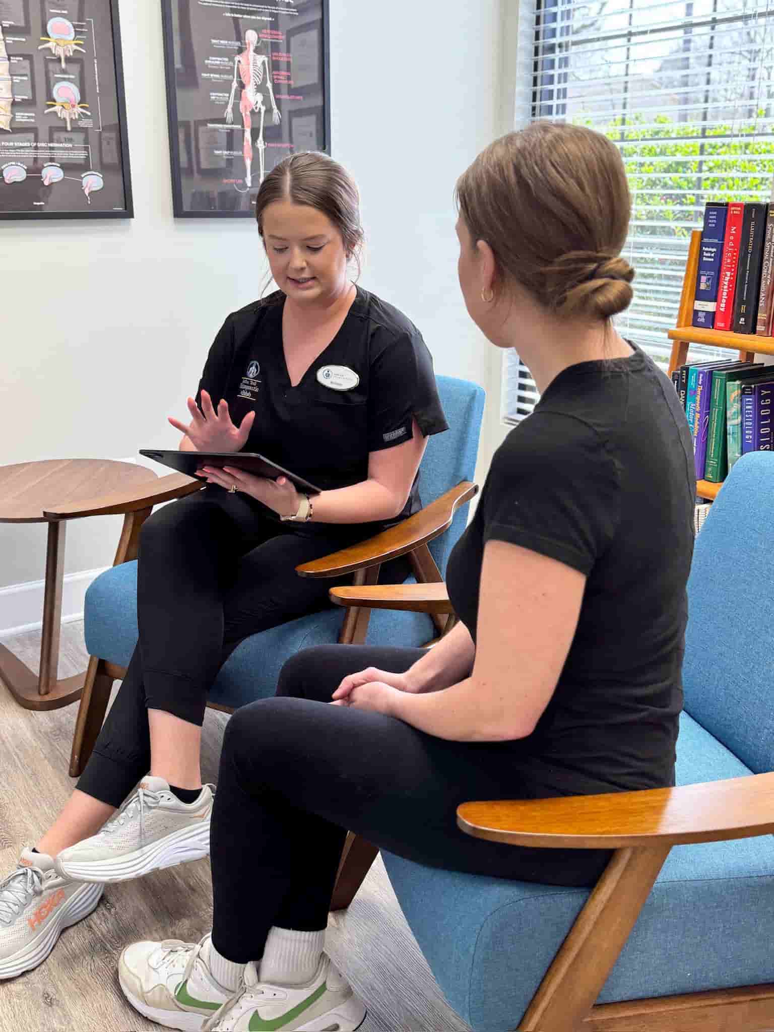 Brittany and a patient sit in armchairs in the waiting area, with Brittany holding a clipboard or tablet as they discuss health history and intake questions.