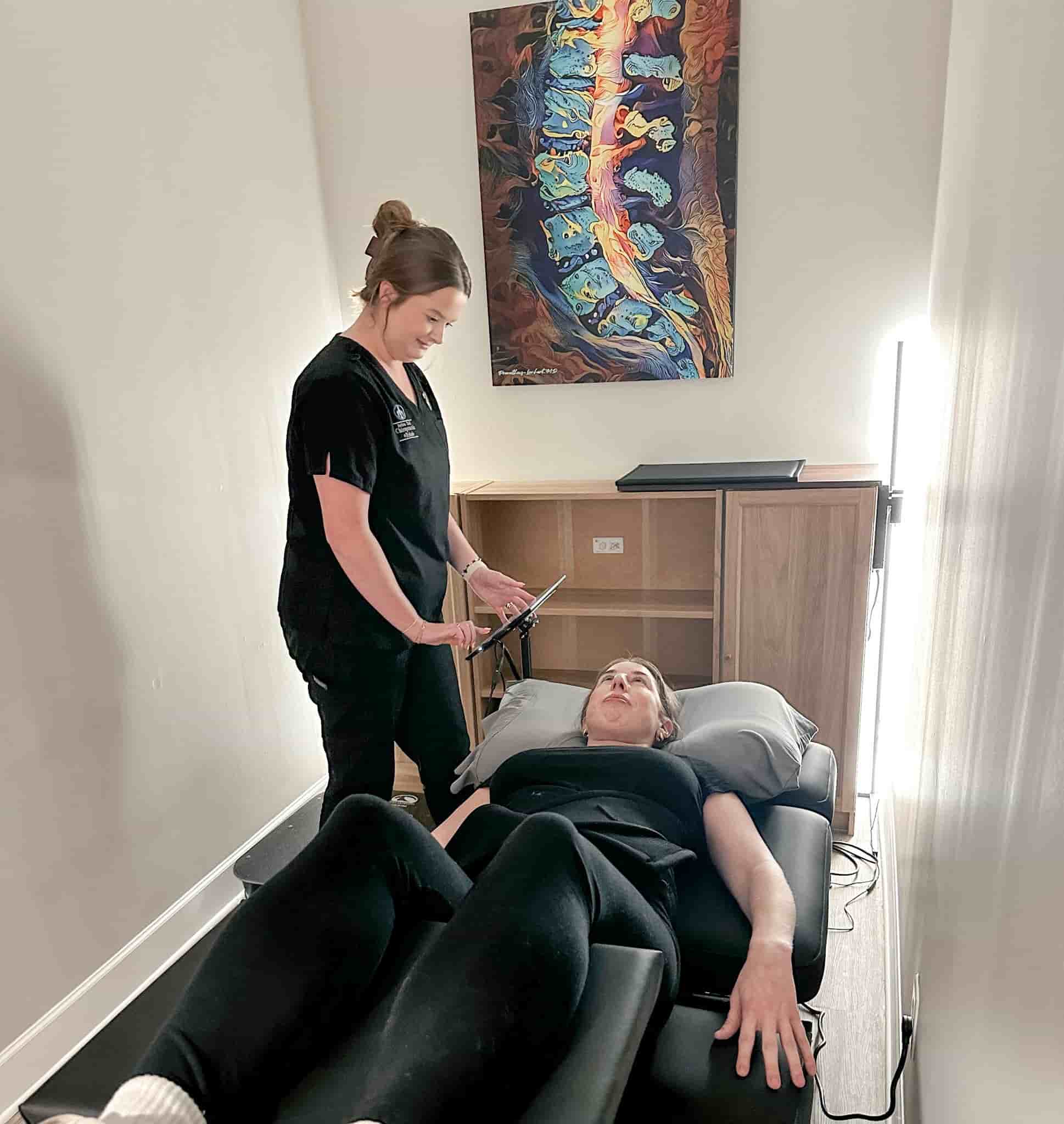 Brittany stands beside a patient lying on a treatment table in a private room, setting up or adjusting therapy equipment near the patient’s legs.