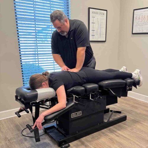 Wide view of Dr. Cameron Gentile delivering a chiropractic adjustment as the patient lies face down on the treatment table in a bright exam room.