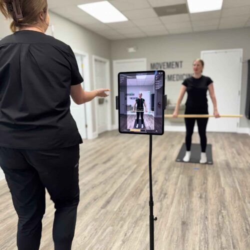 Chiropractic assistant Brittany stands beside a large tablet on a stand while a patient performs a 3D movement screening on a platform.