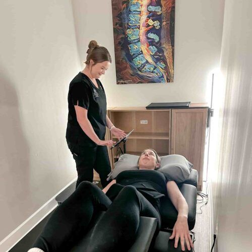 Brittany stands beside a patient lying on a treatment table in a private room, setting up or adjusting therapy equipment near the patient’s legs.