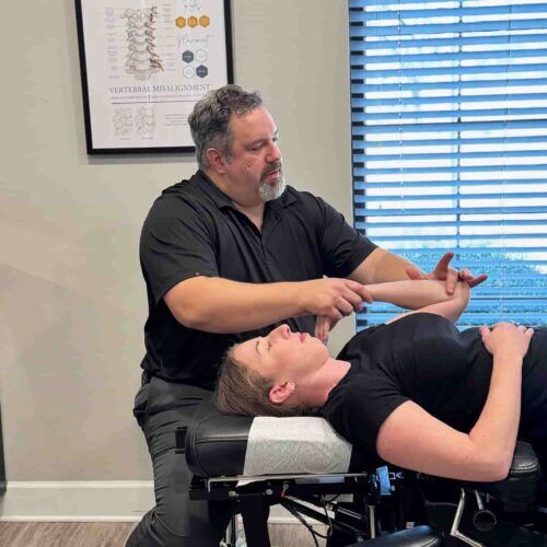 Wide view of Dr. Cameron Gentile seated beside the chiropractic table, adjusting a patient’s neck and upper spine in a bright exam room.