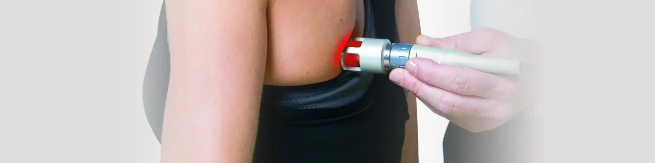 A clinician holds a handheld laser therapy device against a person’s upper arm and shoulder, emitting a red light during a treatment session.
