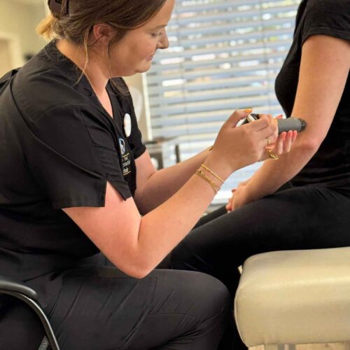Brittany sits beside a patient on the treatment table, holding a handheld therapy device upright and describing how it supports their chiropractic care.