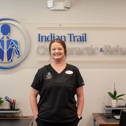 Brittany smiles in front of the Indian Trail Chiropractic & Rehab logo wall at the reception desk, welcoming patients to the clinic.