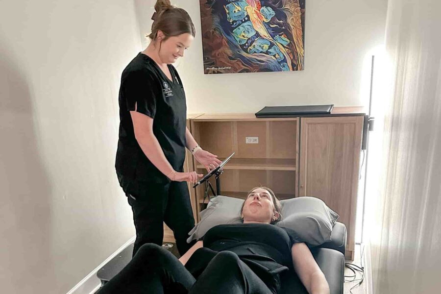 Brittany stands beside a patient lying on a treatment table in a private room, setting up or adjusting therapy equipment near the patient’s legs.