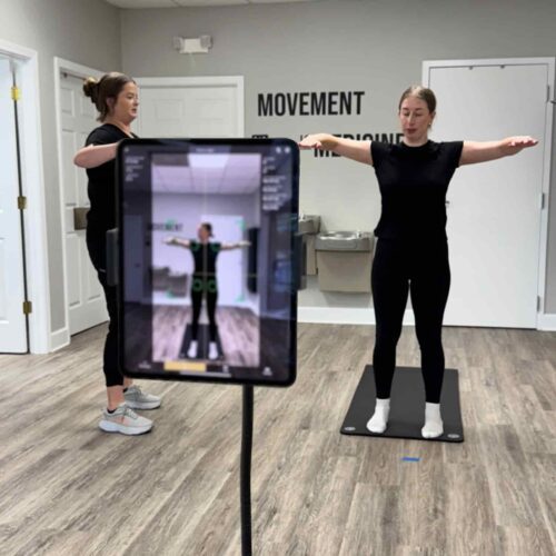 Chiropractic assistant Brittany stands beside a large tablet while a patient holds their arms out on the movement platform during a digital posture and motion screen.