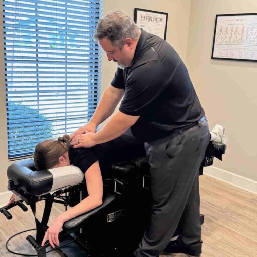 Dr. Cameron Gentile stands over a patient on a drop‑style chiropractic table, applying a thoracic adjustment with his hands on the patient’s lower back.
