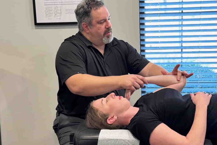Wide view of Dr. Cameron Gentile seated beside the chiropractic table, adjusting a patient’s neck and upper spine in a bright exam room.
