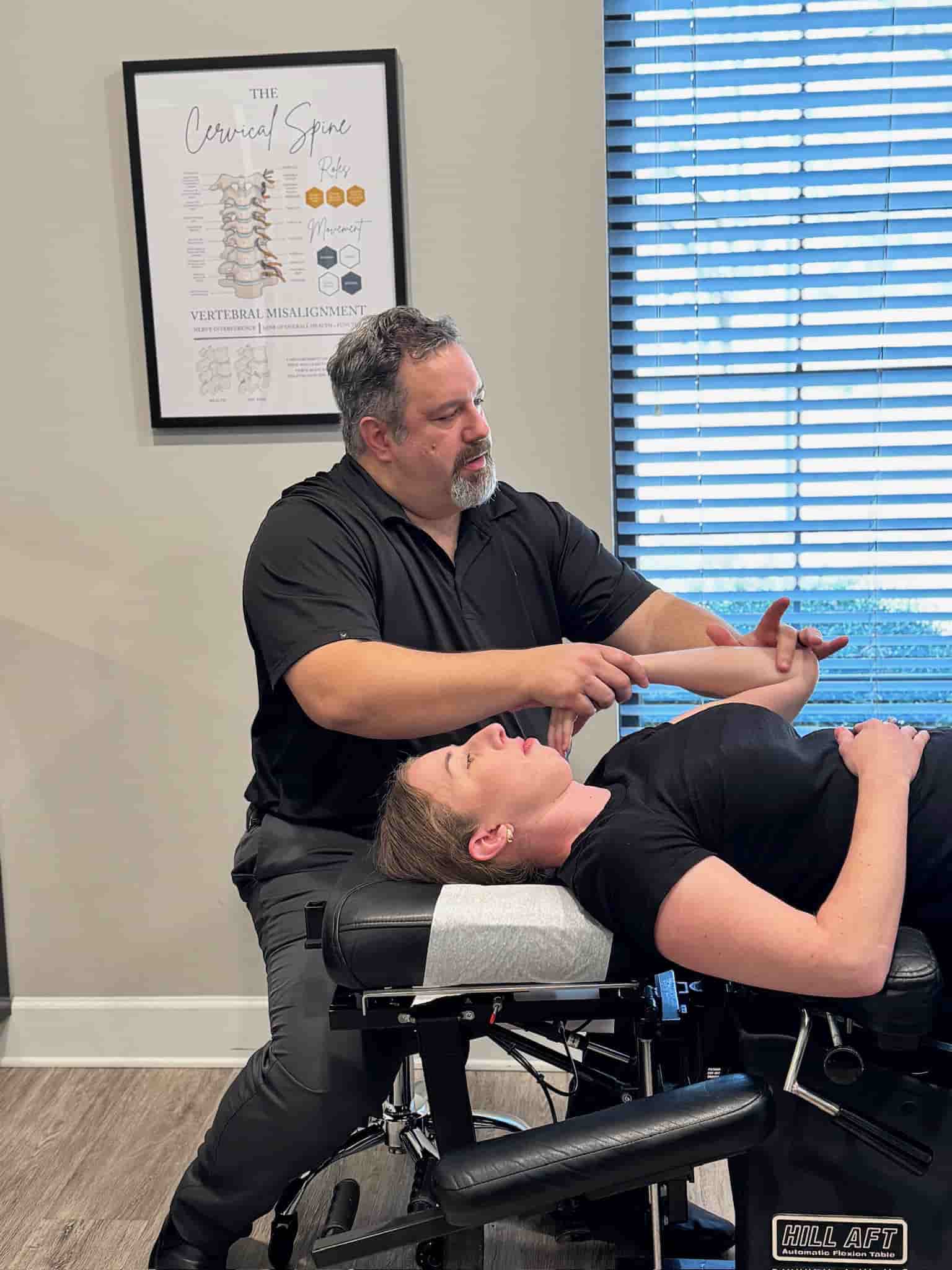Wide view of Dr. Cameron Gentile seated beside the chiropractic table, adjusting a patient’s neck and upper spine in a bright exam room.