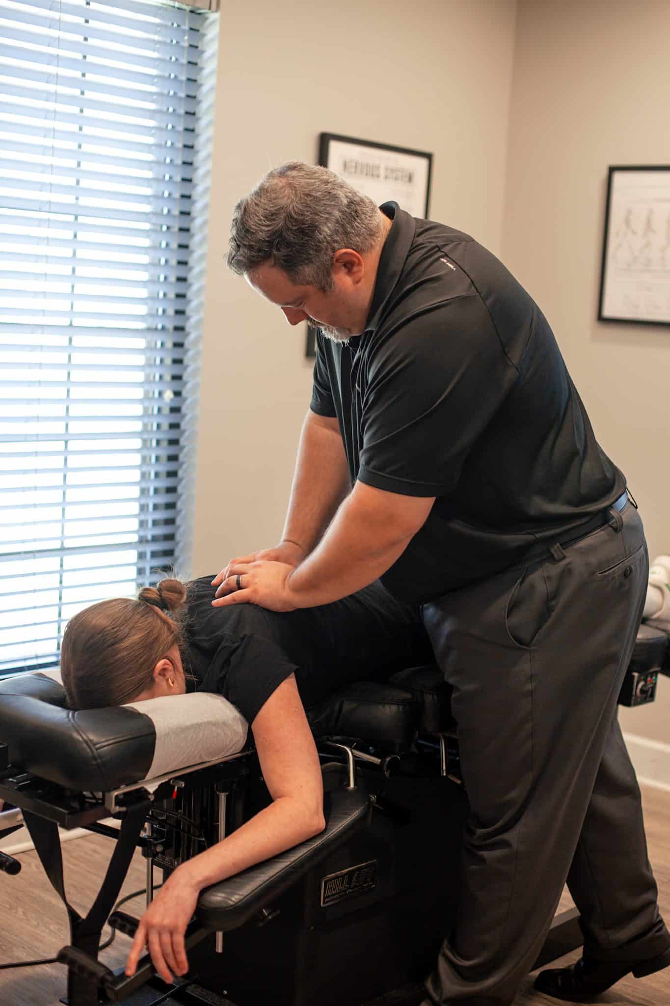 Dr. Cameron Gentile stands beside a chiropractic table, positioning his hands on a patient’s neck and upper back while they lie face down for an adjustment.