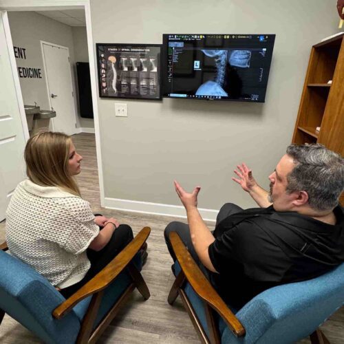 Dr. Cameron Gentile sits beside a patient in chairs facing a wall‑mounted screen, reviewing spinal imaging and movement results together.