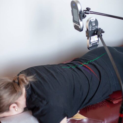 Patient lying face down on a treatment table while a cold laser therapy device with green guide lines is positioned over the lower back.