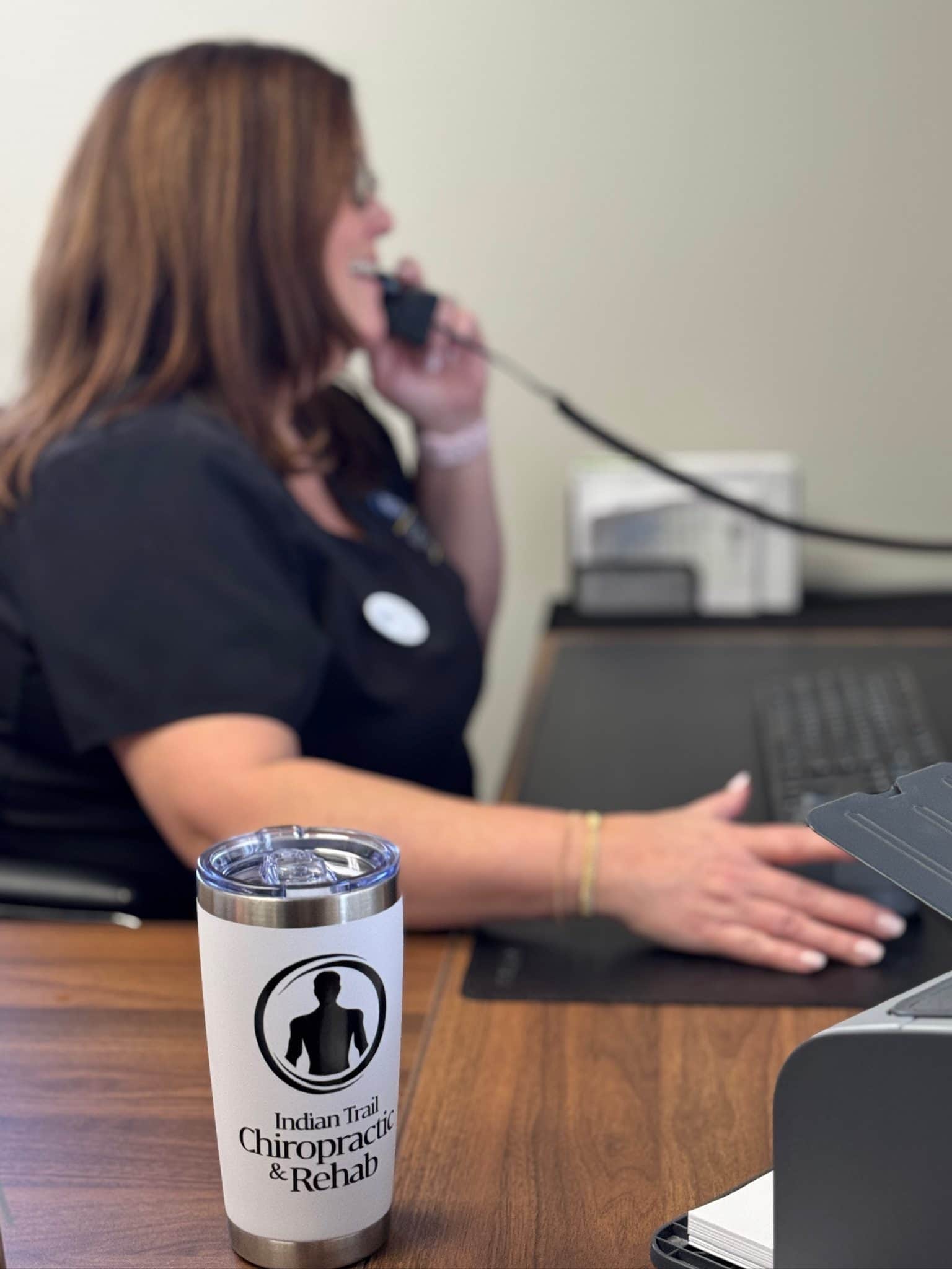 Office Manager Wendy sits at the front desk, talking on the phone while working at the computer, with a branded clinic tumbler on the counter.