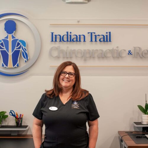 Office Manager Wendy stands smiling in front of the Indian Trail Chiropractic & Rehab logo sign at the front desk.